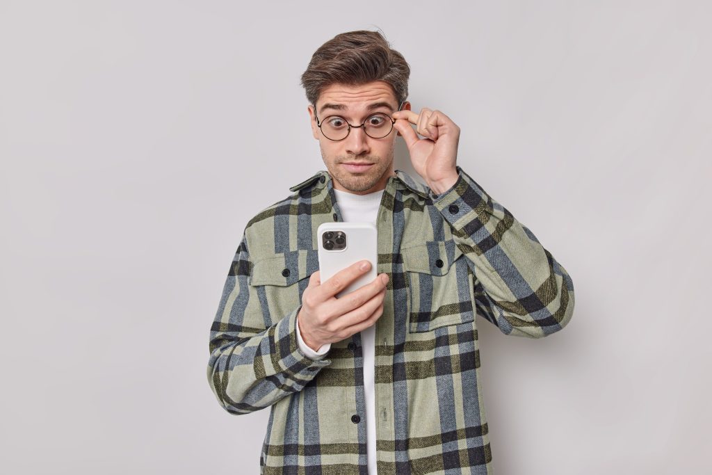 Stunned man keeps hand on rim of spectacles stares at smartphone screen reads shocking news has surprised face wears casual checkered shirt isolated over white background looks at smething amazing